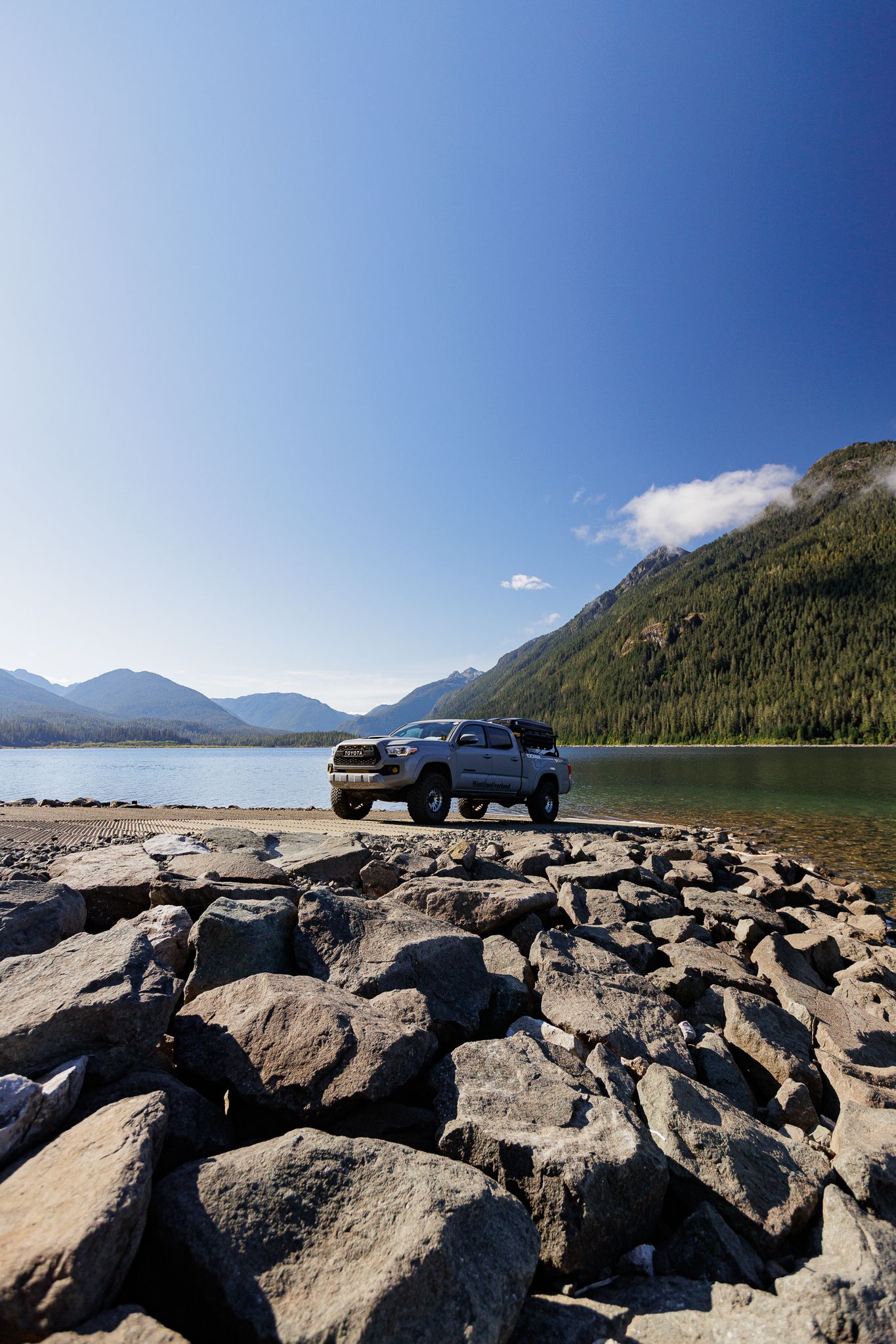 Pickup truck parked on rocky shoreline next to scenic mountain lake with forested peaks and blue skies.