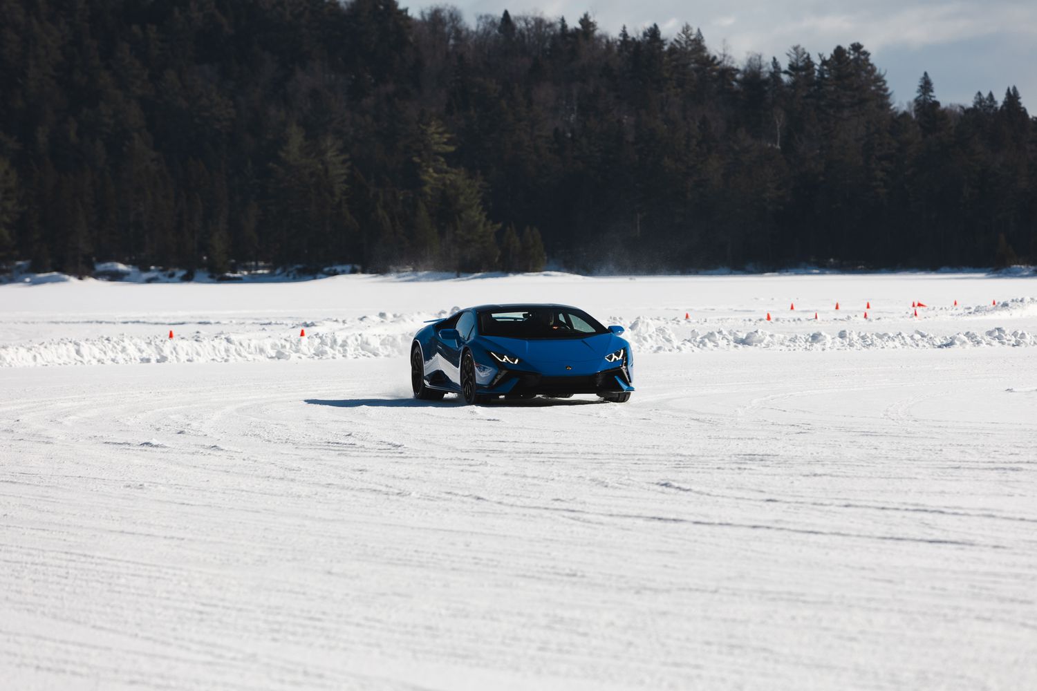 A blue car drives across a vast snowy lake surface with evergreen trees visible in the background.