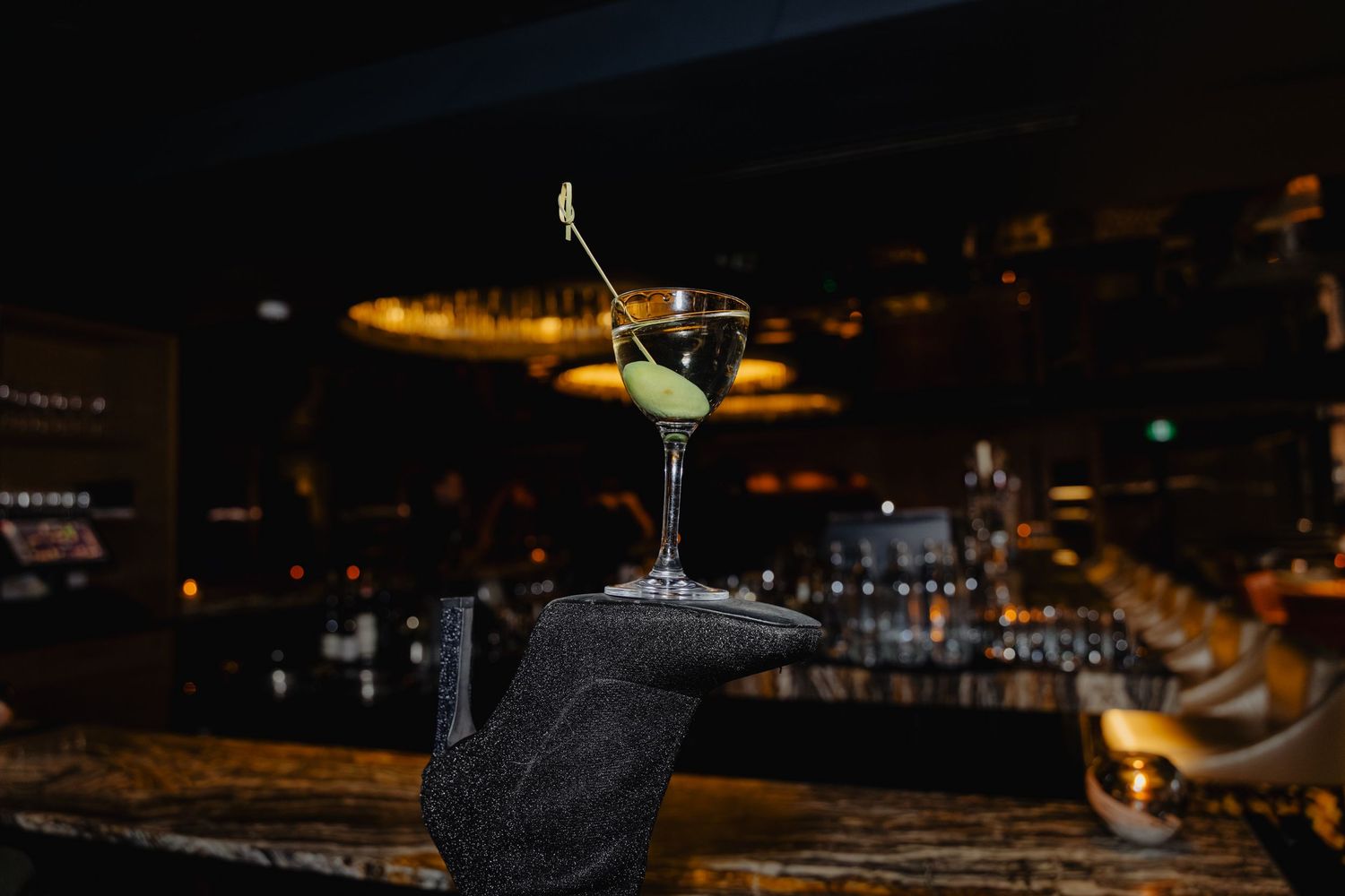 A row of martini glasses with cocktails sit on a dark bar counter illuminated by warm ambient lighting.