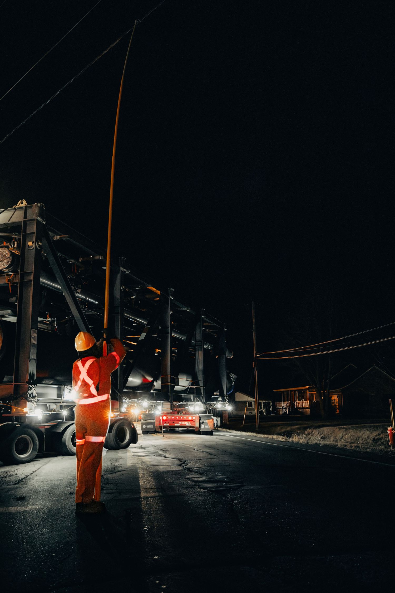Utility worker in orange safety gear performs maintenance on power lines at night with trucks and equipment visible.