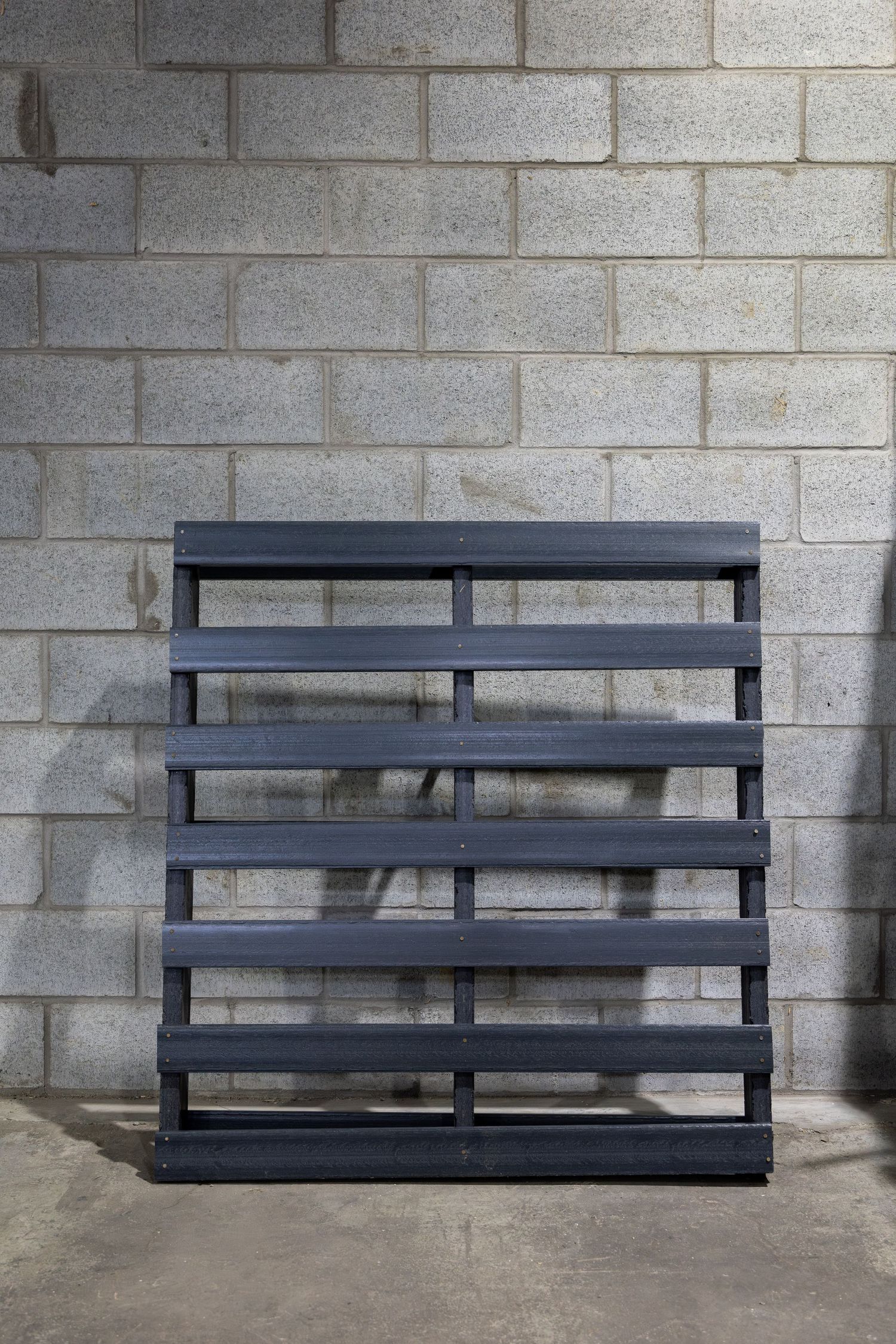 Black painted wooden pallet stands against concrete block wall in industrial setting.