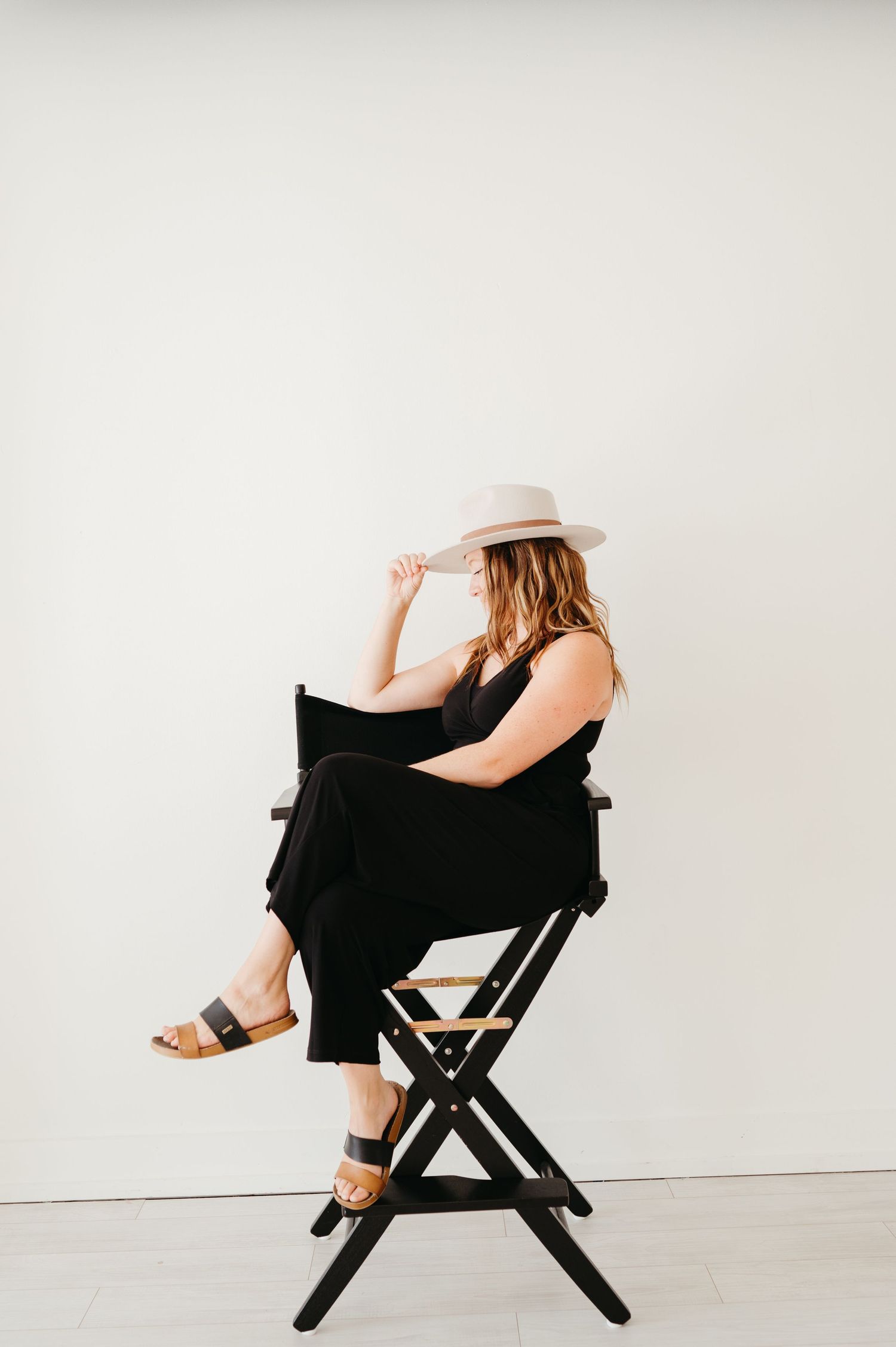 Woman in black dress hat sits elegantly on director's chair against white background.