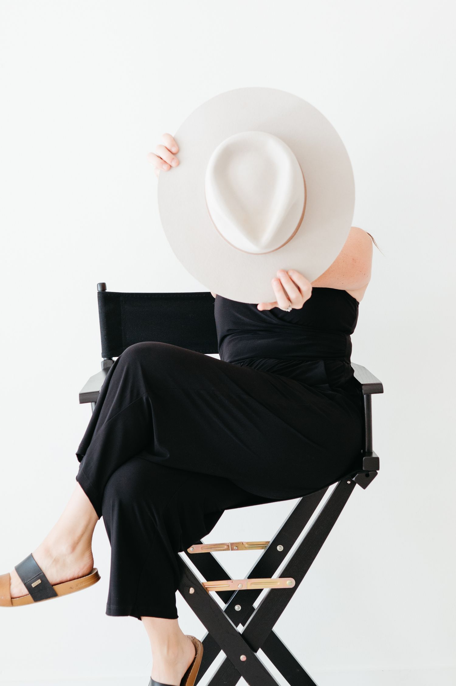 A person in black attire sits on a director's chair holding a white wide-brimmed hat against a white background.