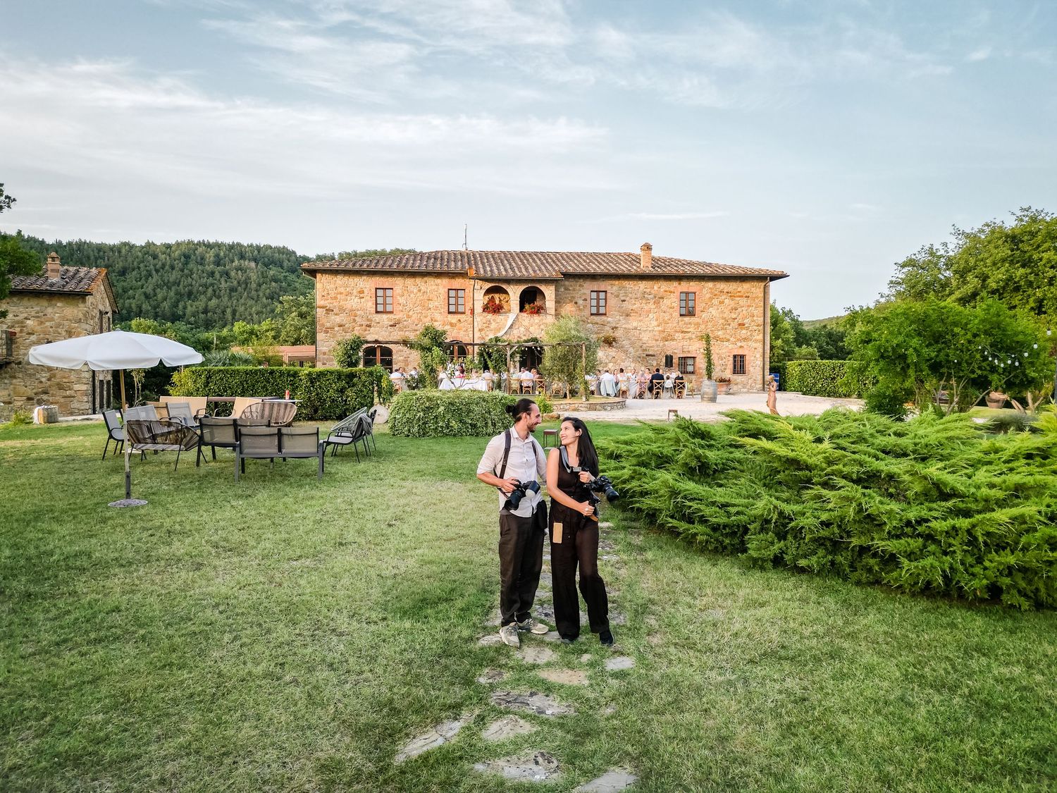 Two tourists explore lush gardens of a rustic Tuscan stone villa surrounded by cypress trees in Italy.