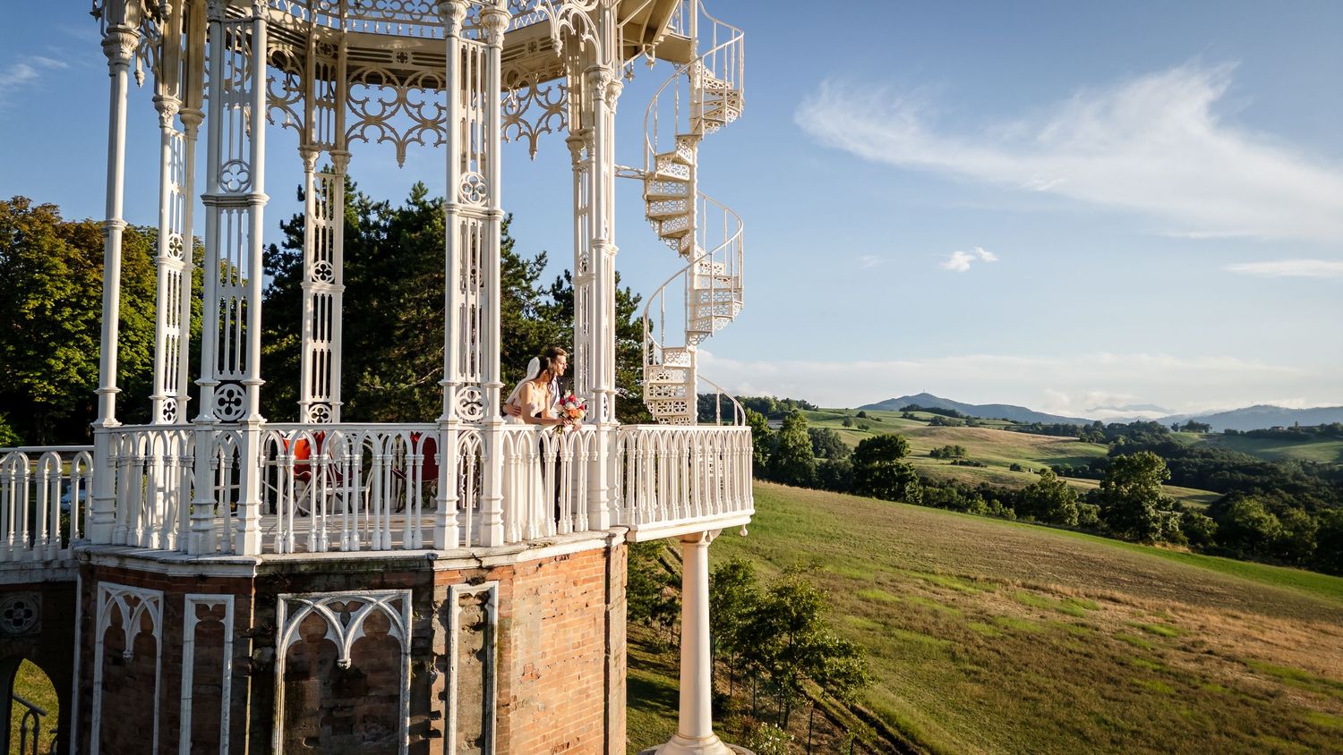 Ornate white Victorian gazebo on brick base overlooking rolling green hills under blue sky.