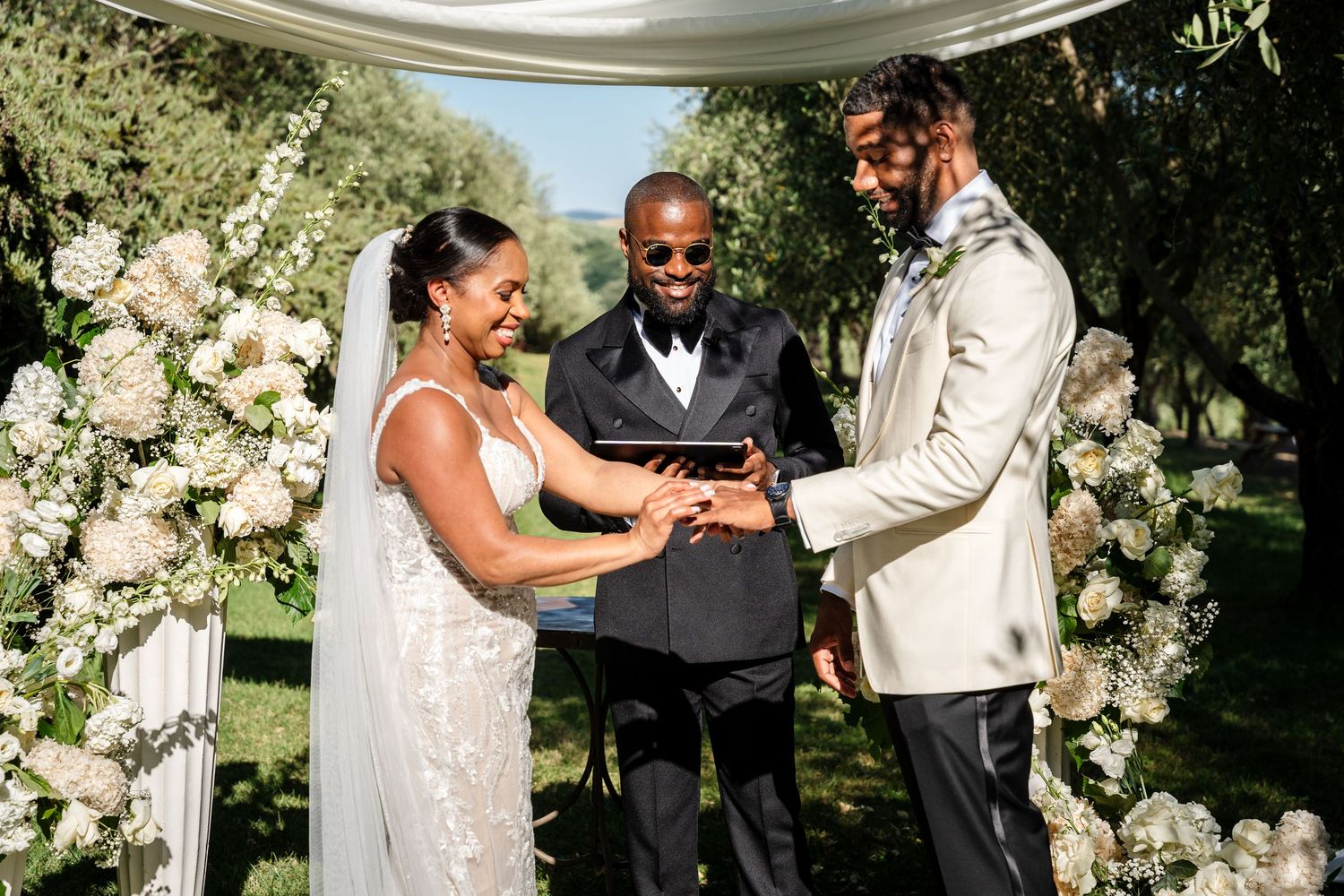 Bride and groom exchange rings outdoors at floral arch wedding ceremony with smiling officiant.