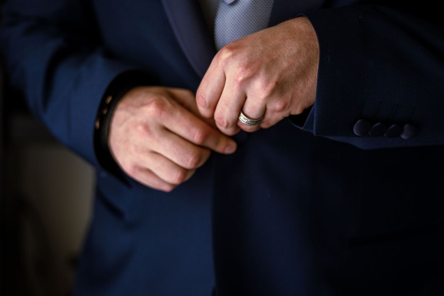 Man in navy blue suit adjusting jacket buttons, wearing a silver wedding ring, close-up detail shot.
