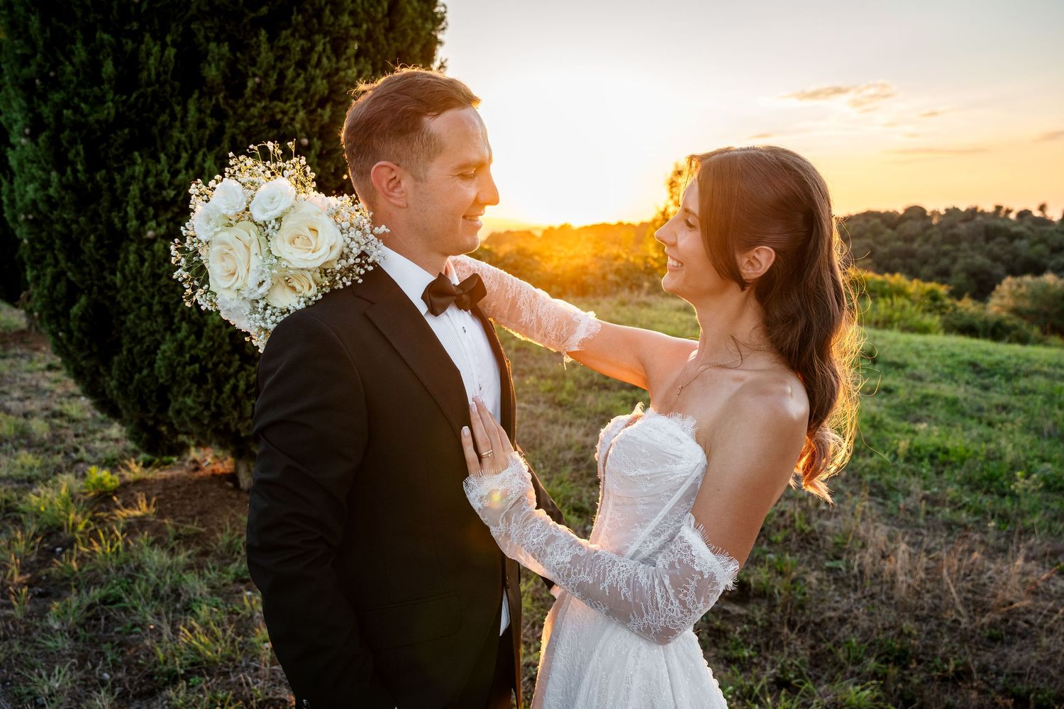 Bride and groom share a romantic sunset moment outdoors, with the bride holding a white bouquet and touching the groom's face.