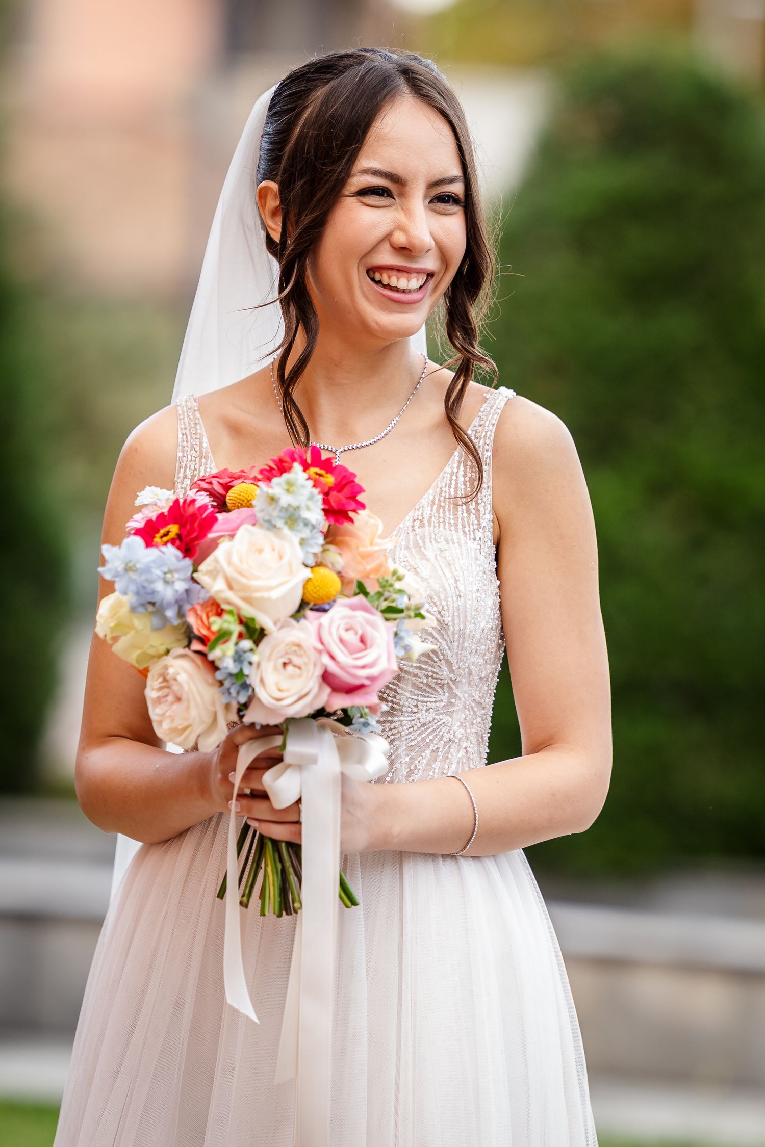 Joyful bride in beaded gown holding colorful wedding bouquet with roses and wildflowers outdoors.