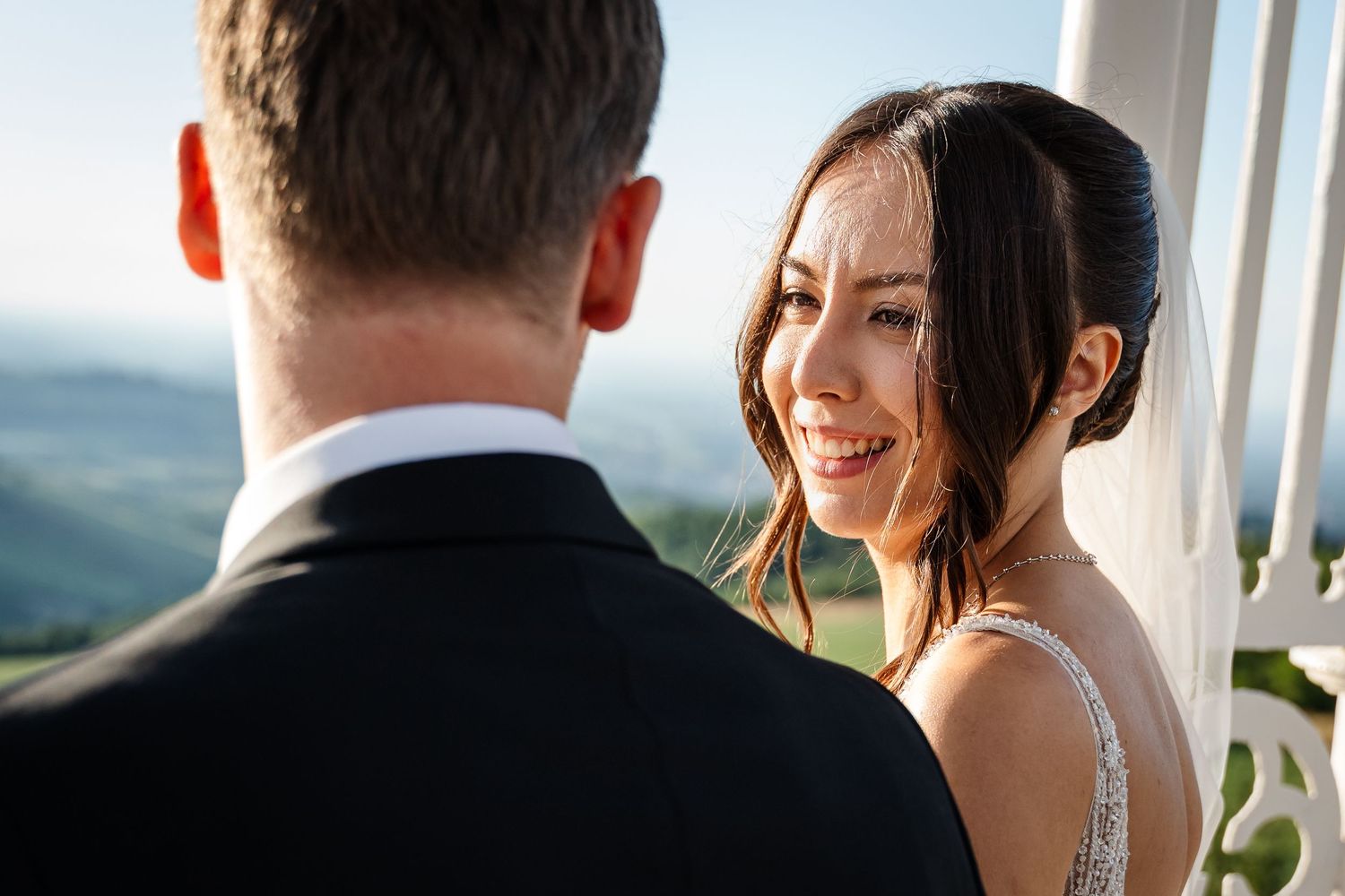 Bride smiling joyfully at groom during outdoor wedding ceremony with scenic green hills in the background.