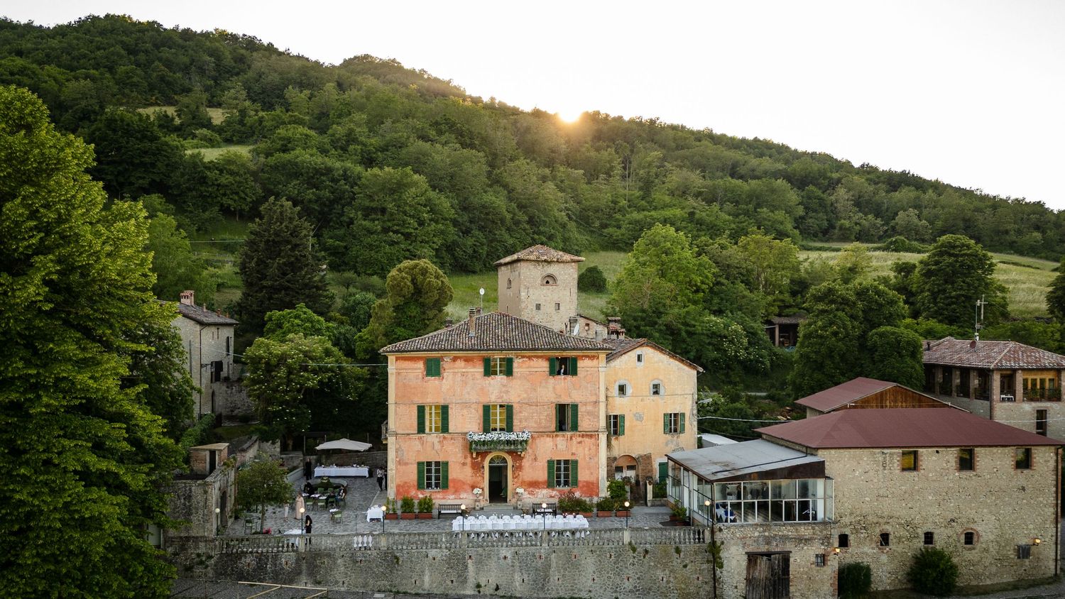 Aerial view of a rustic Italian villa with peach facade nestled among lush green hills at golden sunset.