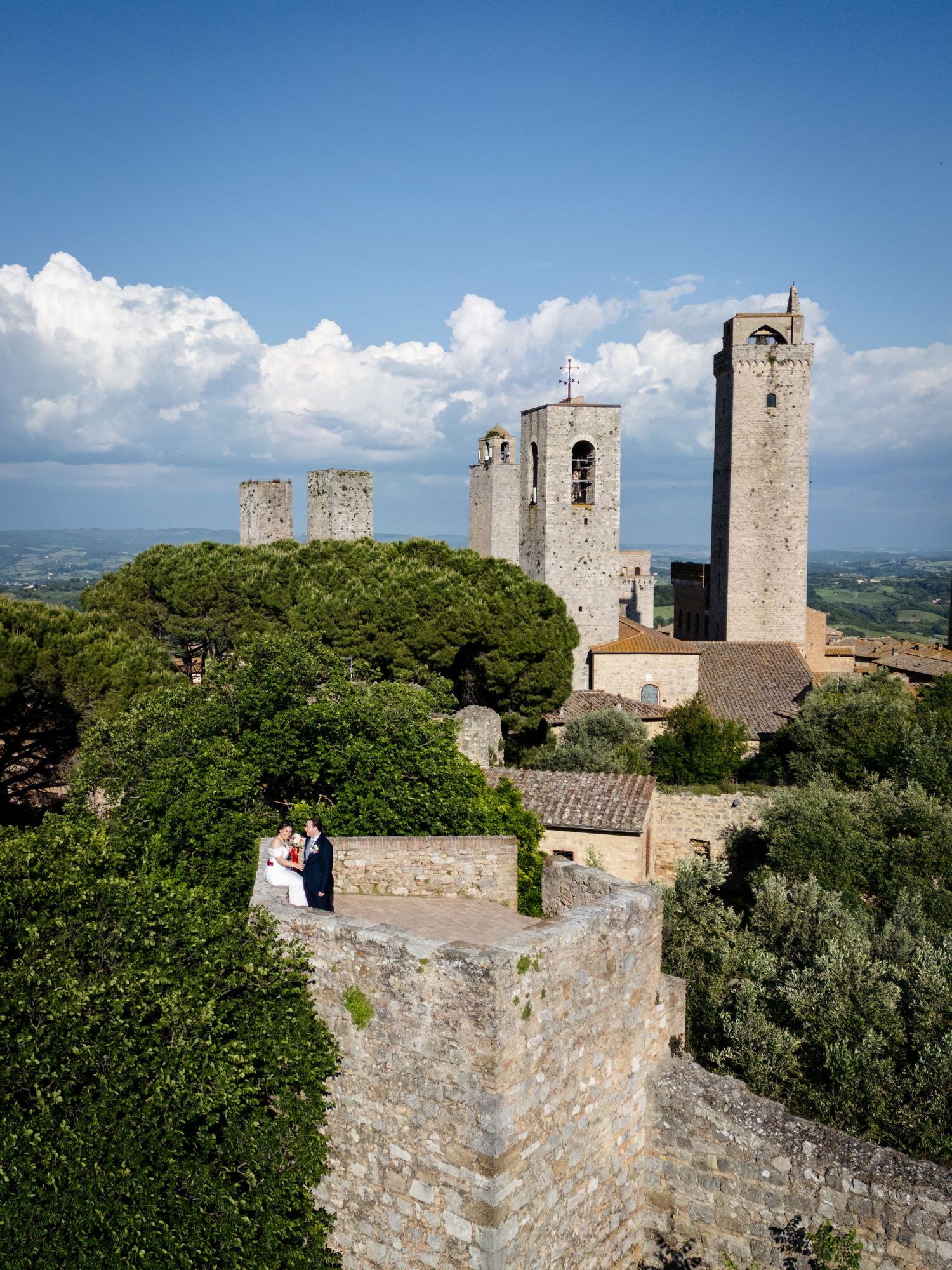 Couple poses on ancient stone walls of San Gimignano with medieval towers and Tuscan landscape backdrop.