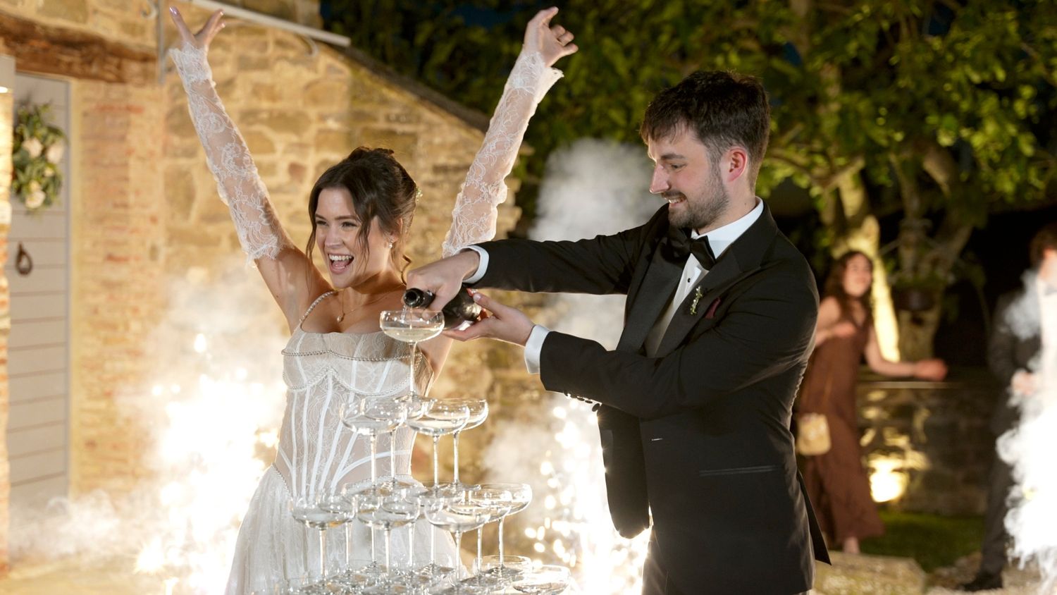 Bride and groom pouring champagne tower at elegant outdoor wedding reception with sparkling fireworks.