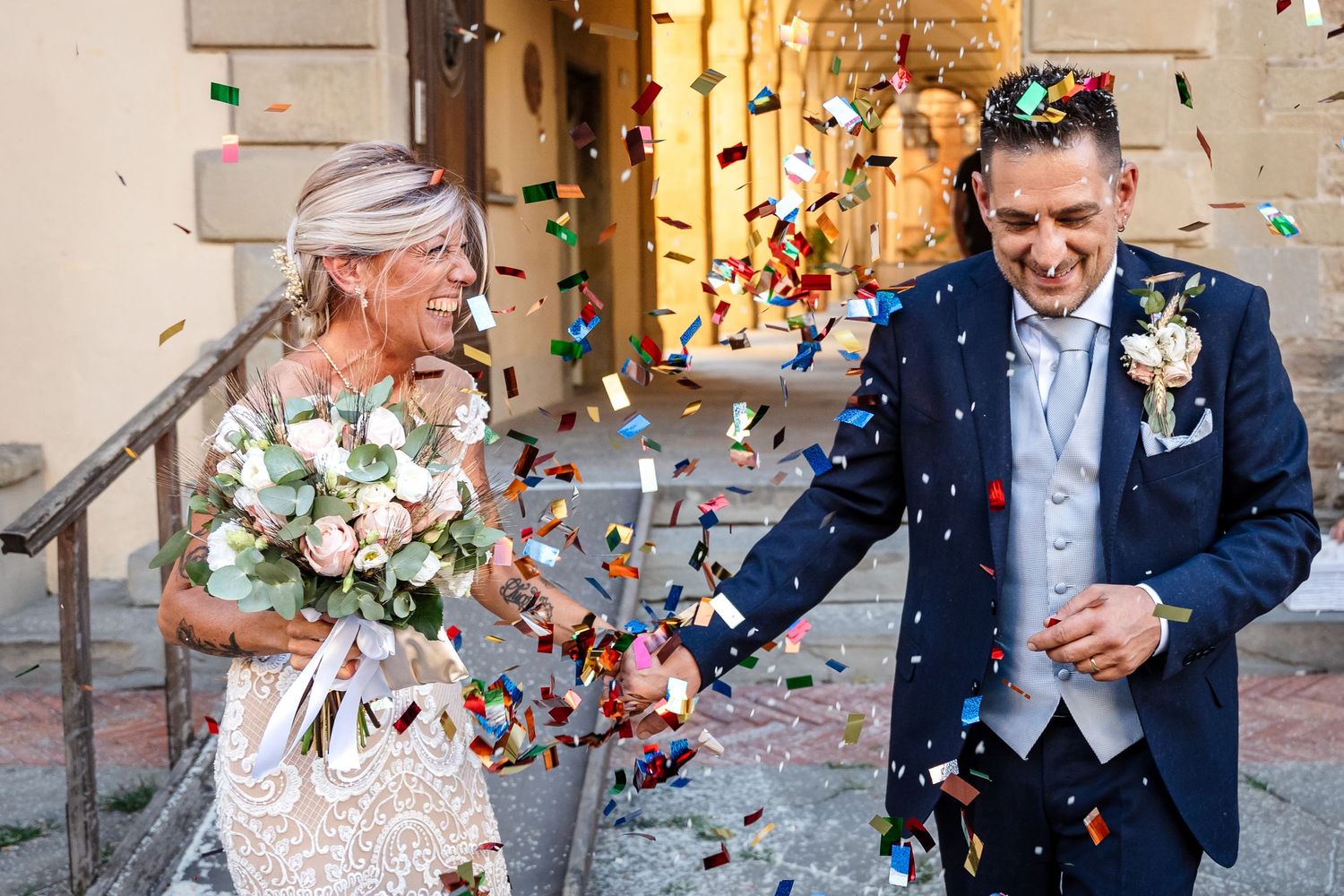 Wedding guests throw pink and blue confetti over the newlyweds during a joyful outdoor celebration.