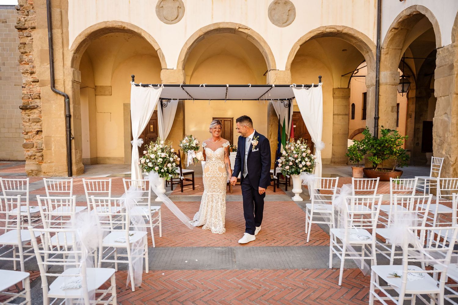 Wedding ceremony setup with white chairs arranged in rows under elegant stone archways in a Mediterranean-style courtyard.