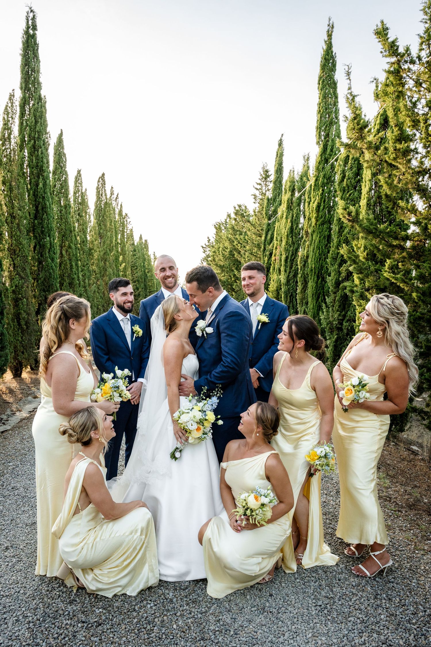 Bride and groom kiss surrounded by bridesmaids in yellow dresses and groomsmen in navy suits outdoors with cypress trees.