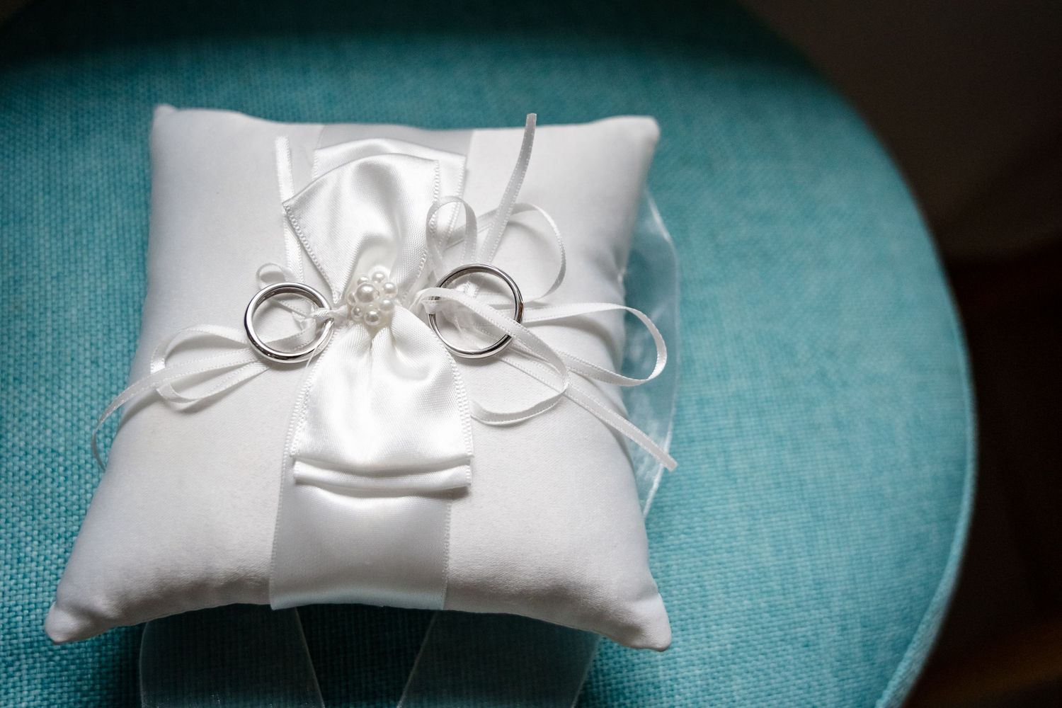 Two silver wedding rings tied with white ribbon on a white satin ring bearer pillow.