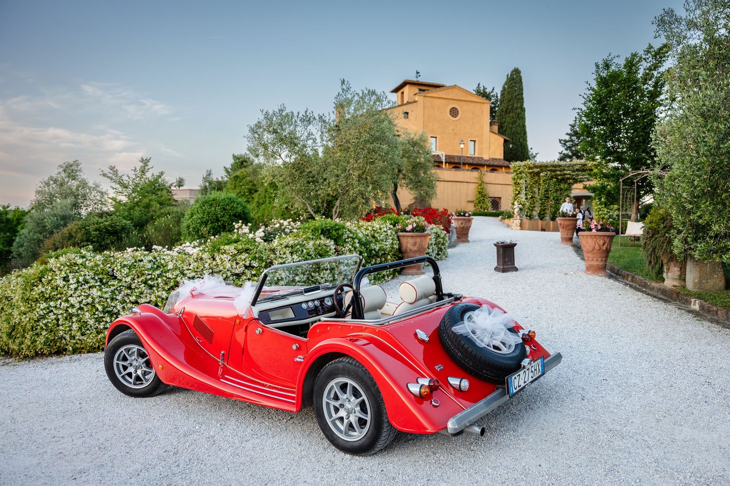 A vintage red convertible sports car parked on a gravel driveway in front of an Italian villa with gardens.