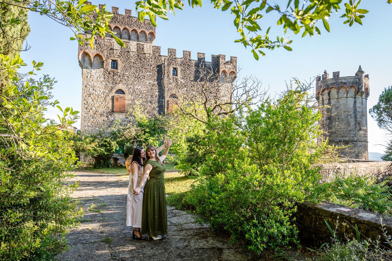 Two women in elegant dresses pose before a historic stone castle surrounded by lush green gardens in Italy.