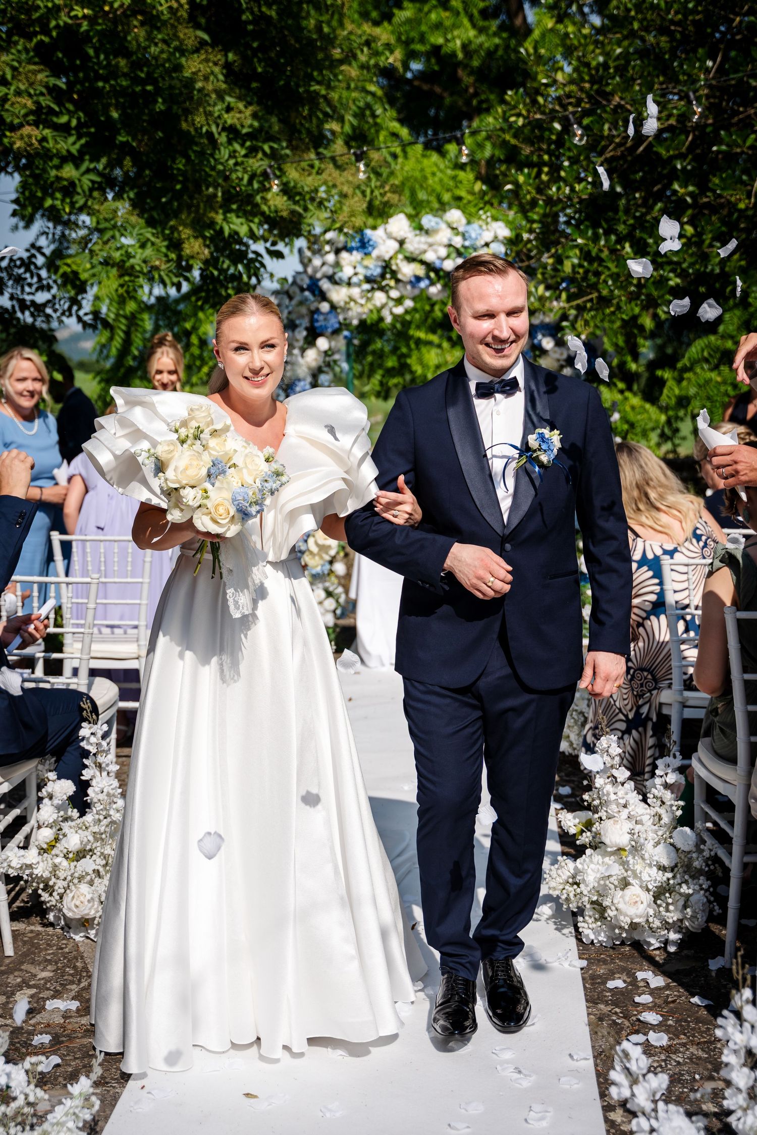 Bride and groom walk down outdoor wedding aisle, bride in white ruffled gown holding bouquet, groom in navy tuxedo.
