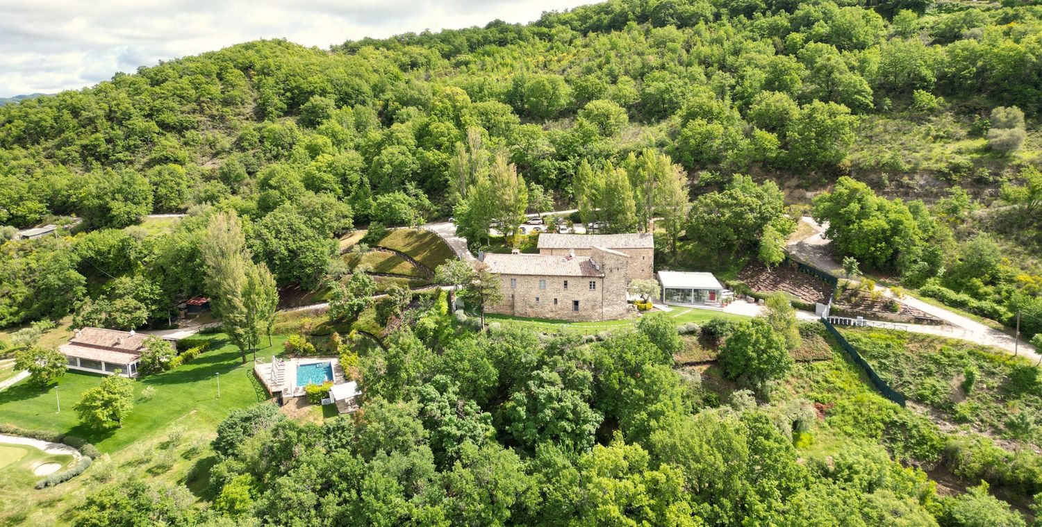 Aerial view of a stone farmhouse surrounded by lush green trees with a swimming pool and gardens in a rural French countryside setting.