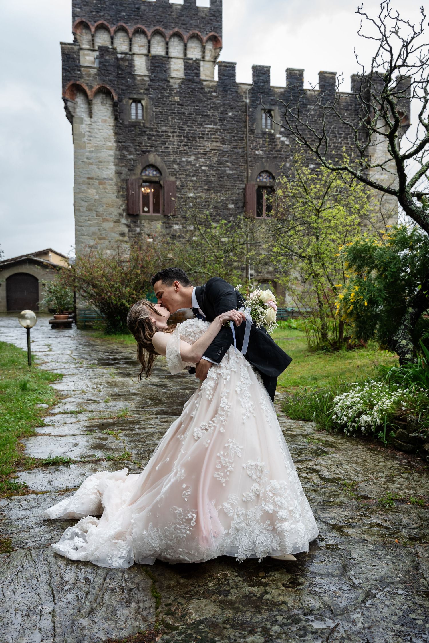 A romantic wedding couple embraces in front of a medieval stone castle tower on a rainy day in Italy.