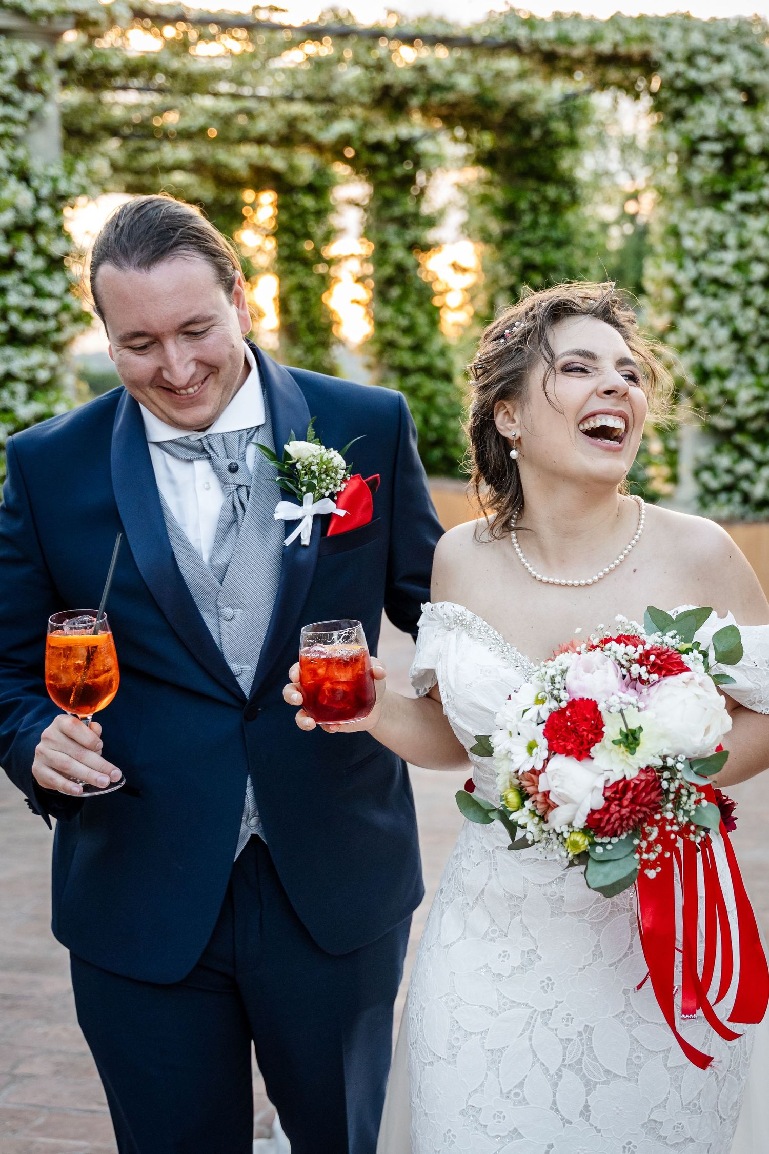 Laughing bride and groom walking together outdoors, holding Aperol Spritz cocktails, bride carrying red and white bouquet.