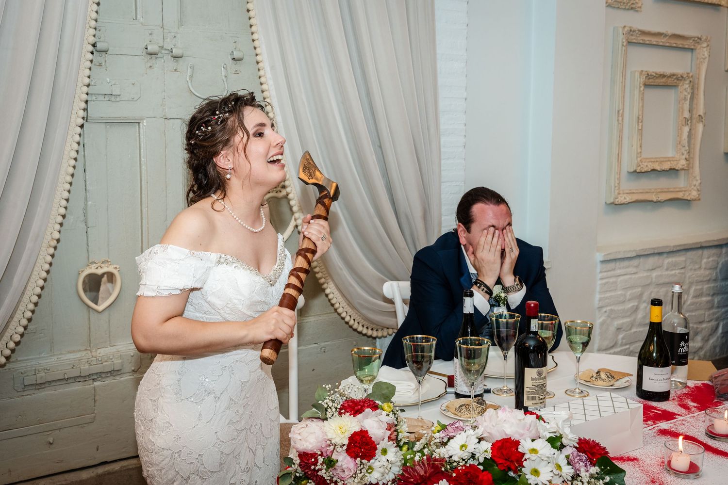 Bride in lace gown holds decorative scepter while groom laughs at wedding reception table.