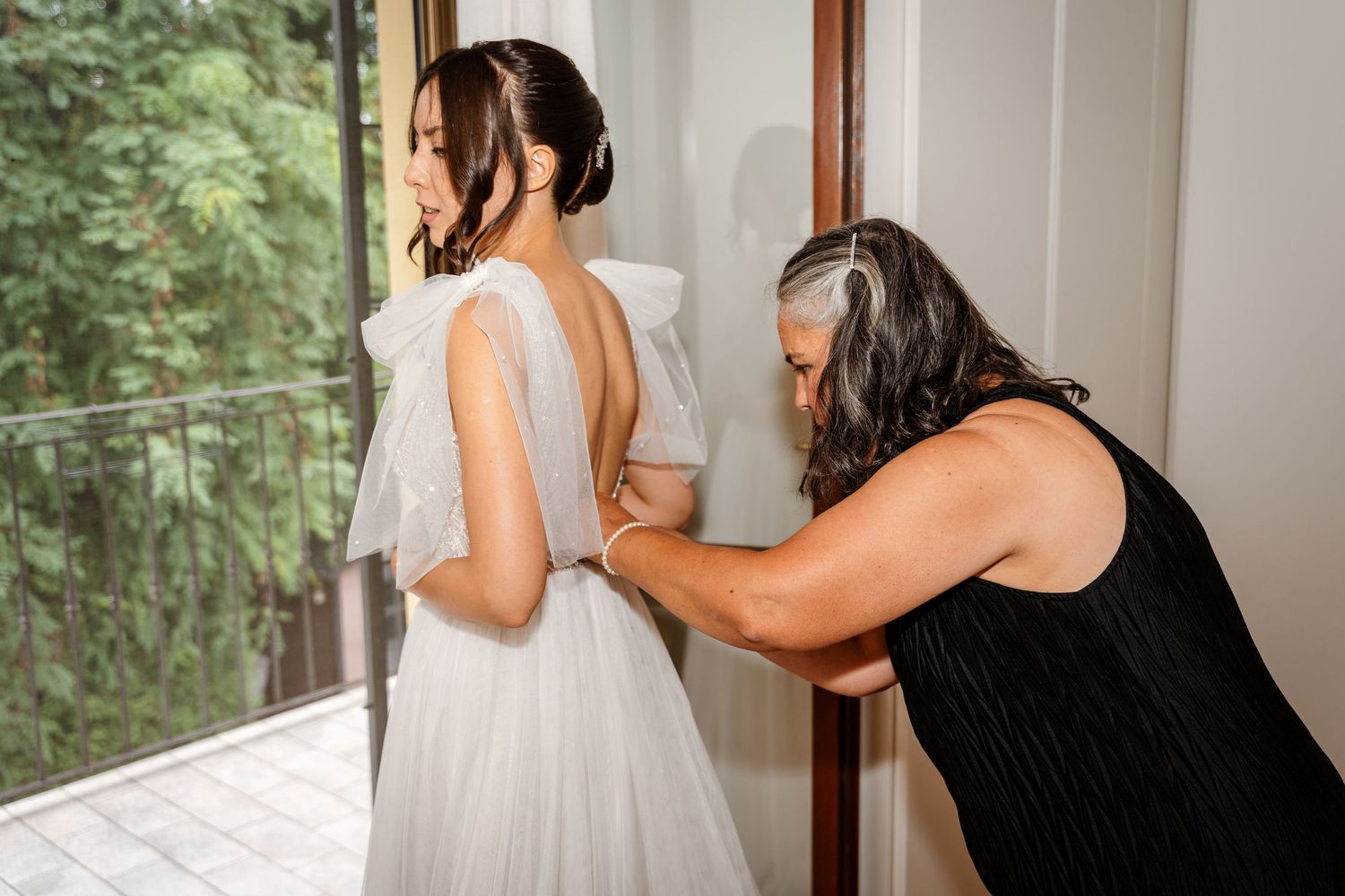 Mother helping bride button up elegant white wedding dress near window.