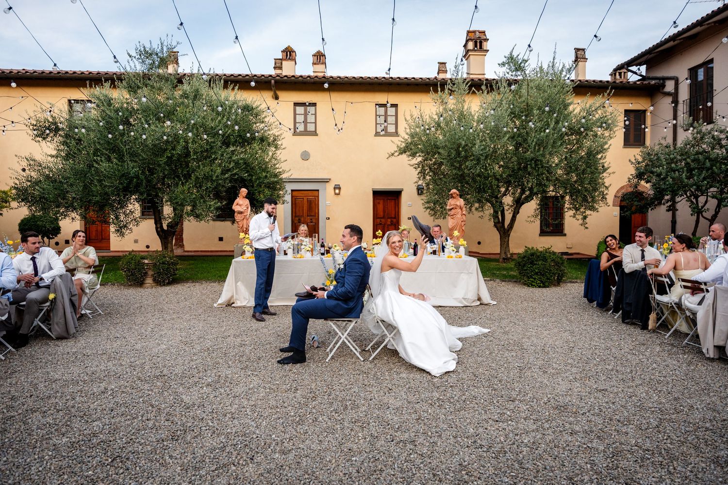Outdoor wedding reception in a rustic Italian courtyard with olive trees, string lights, and guests seated at elegant tables.