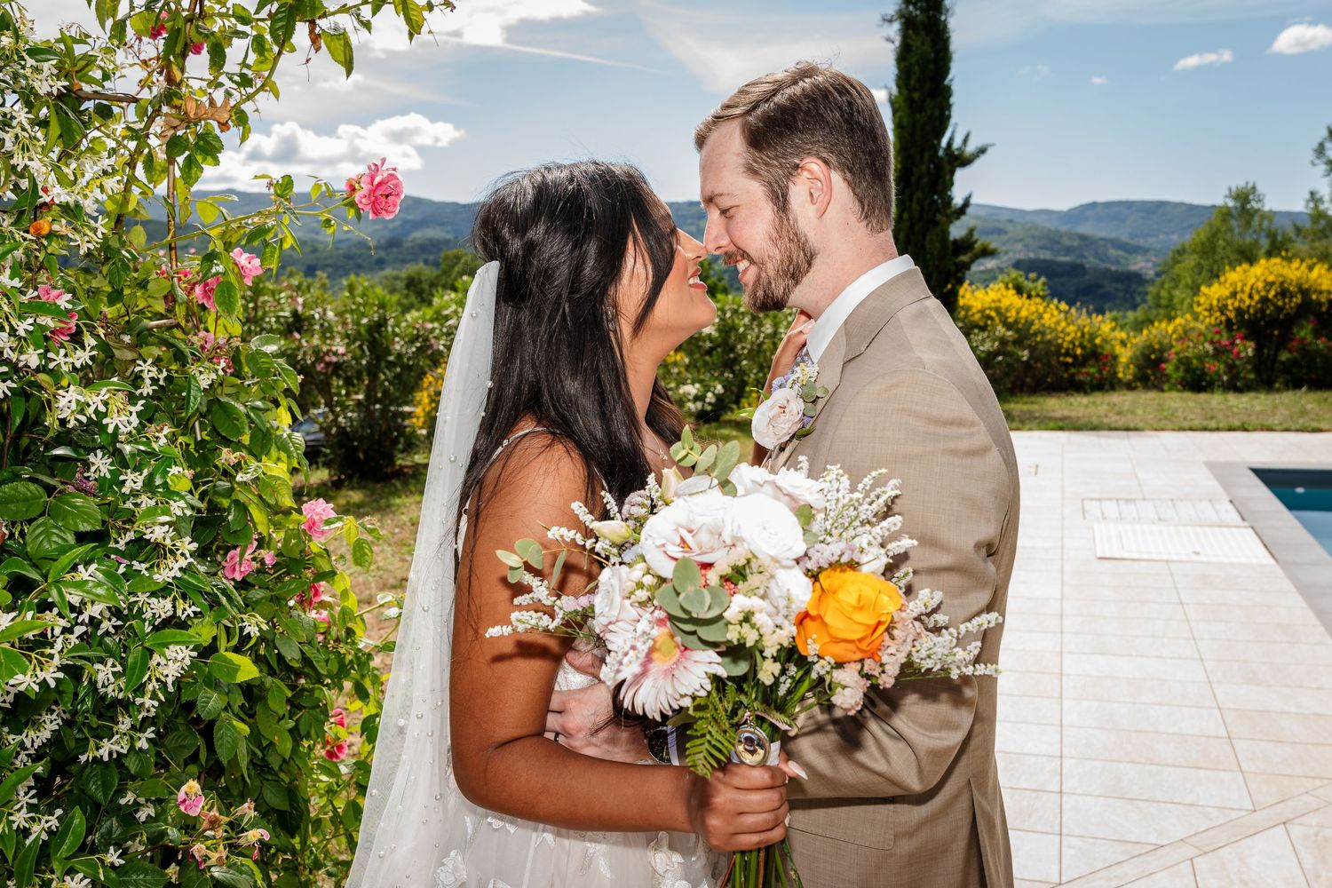 Bride and groom share a romantic moment, holding a colorful bouquet, with lush greenery and mountains in the background.