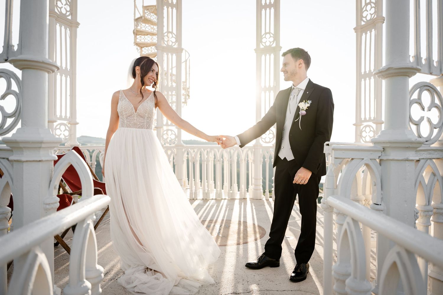 Bride and groom holding hands on a sunlit white balcony, bride in flowing gown, groom in black tuxedo.