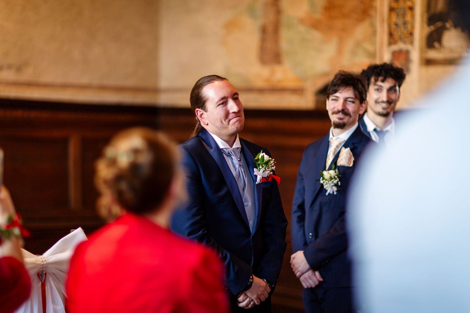Groom smiles emotionally as bride approaches altar, groomsmen standing beside him in navy suits.