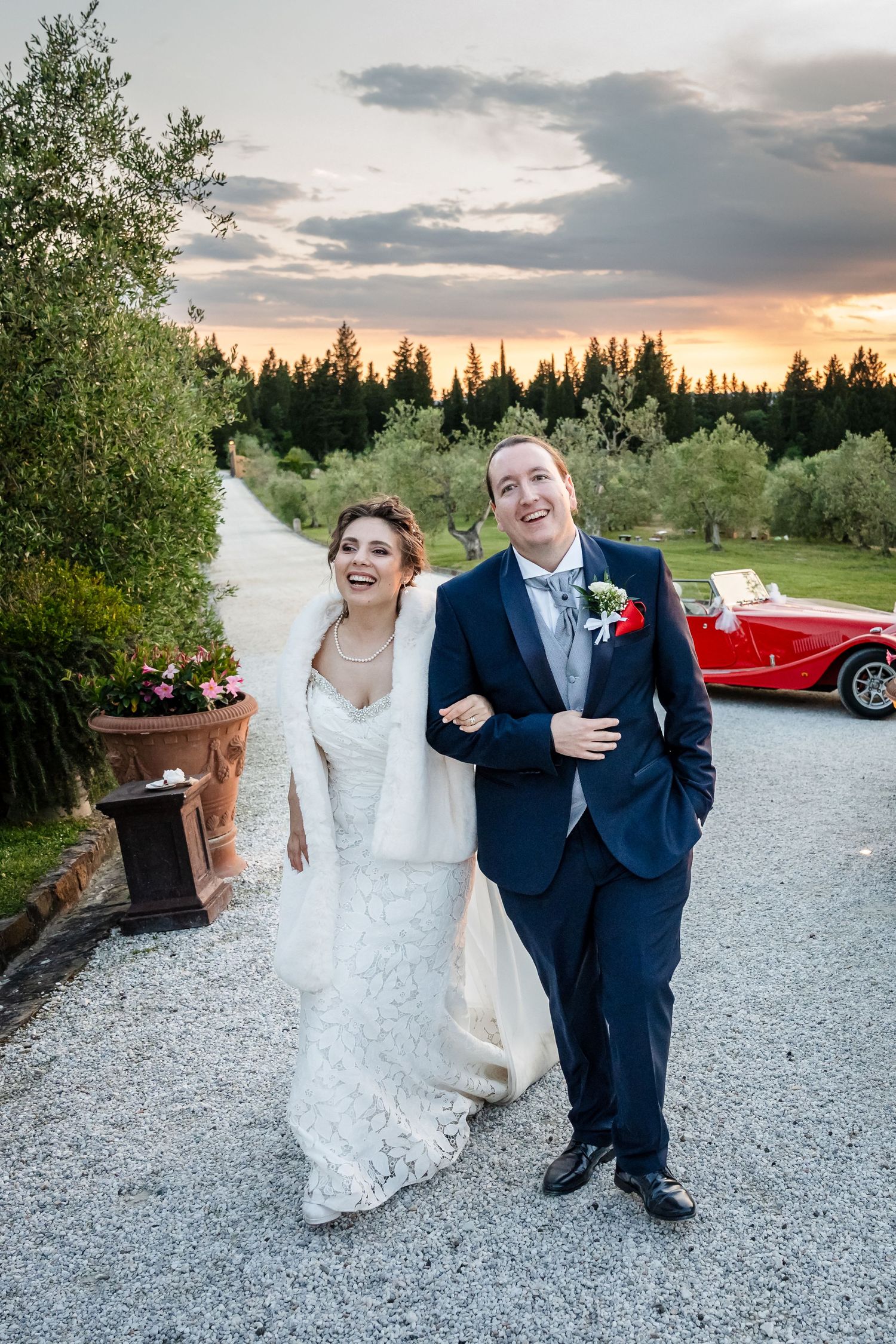 A joyful bride and groom walk hand in hand on a gravel path at sunset, with a red vintage car and trees in the background.
