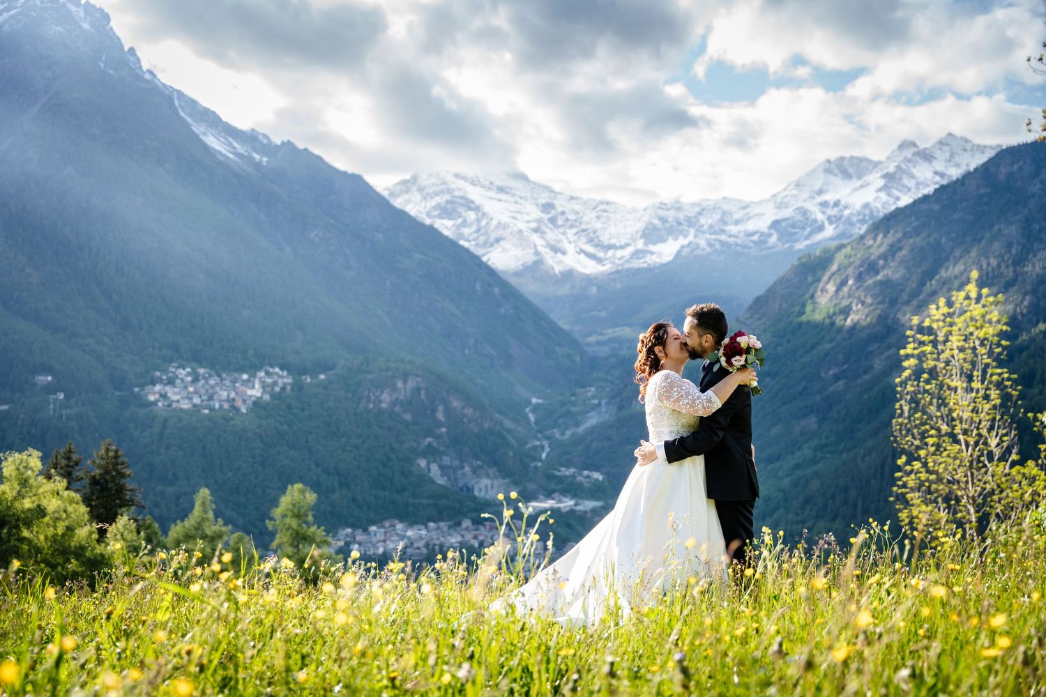 A couple in wedding attire embraces in a scenic mountain meadow with snow-capped peaks in the background.