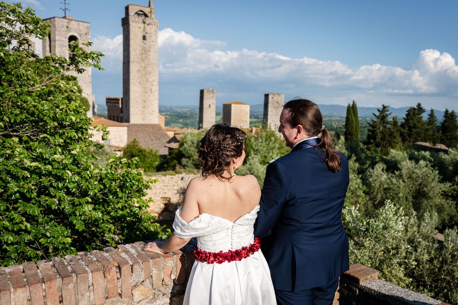 A couple overlooks the scenic Tuscan countryside with stone buildings and cypress trees against a blue summer sky.