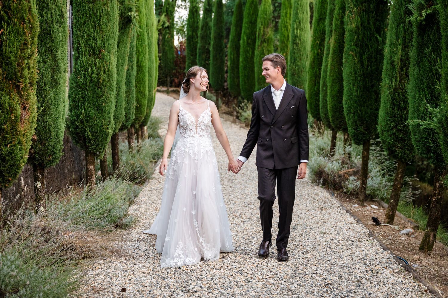 Bride and groom walk hand-in-hand along a gravel path lined with tall cypress trees on their wedding day.