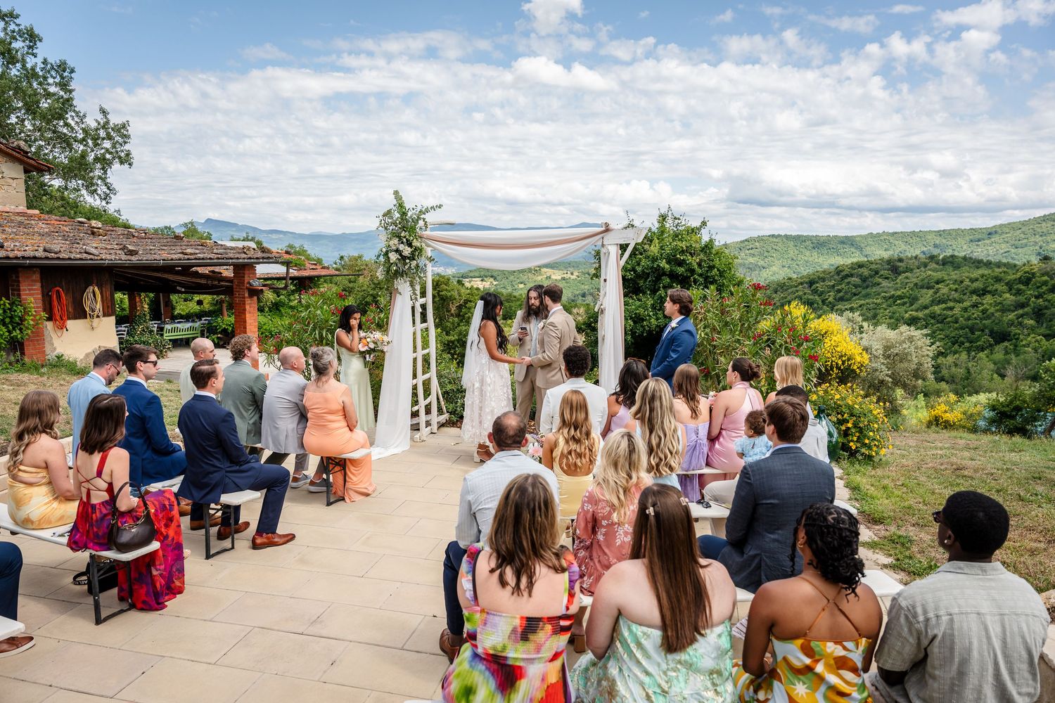 Outdoor wedding ceremony on a terrace with rolling green hills backdrop, guests seated as couple exchanges vows under floral arch.