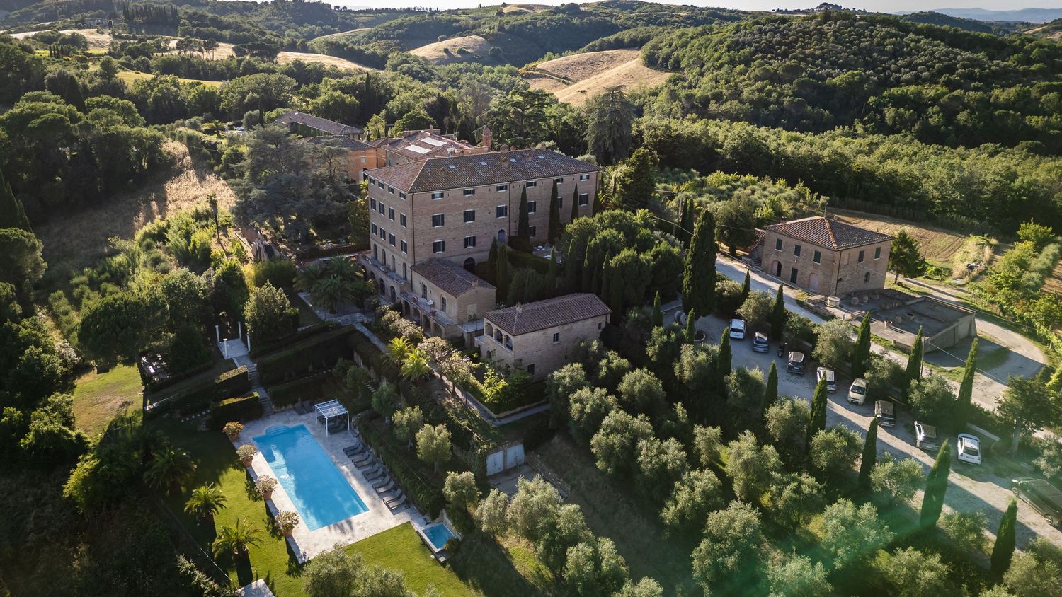 Aerial view of a historic Tuscan villa with a swimming pool surrounded by cypress trees and rolling hills.