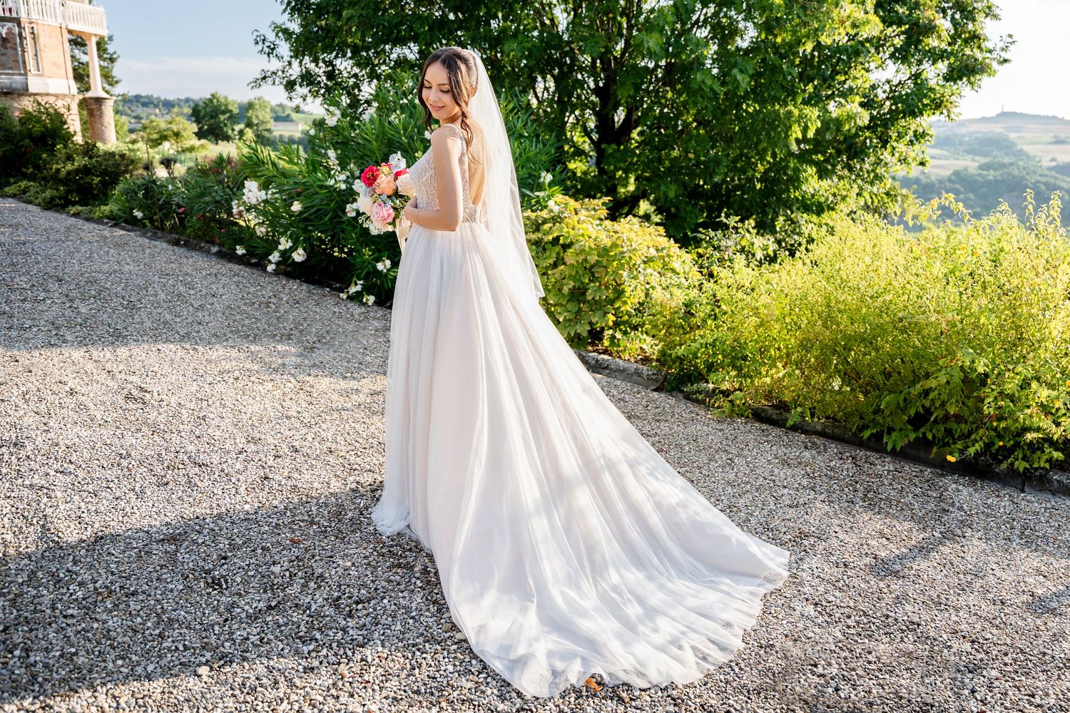 Bride in elegant white gown with long train holding bouquet on gravel path surrounded by lush greenery.