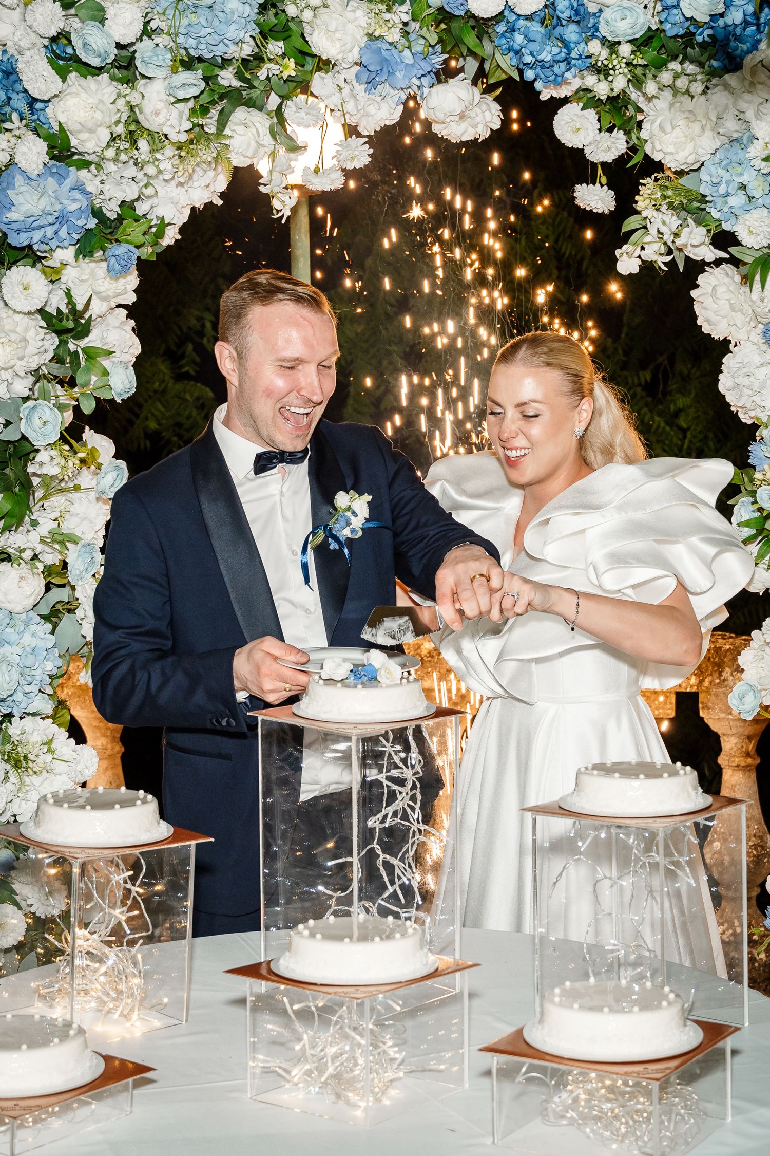 A couple in formal attire cuts their elegant white wedding cake decorated with flowers and sparklers at their reception.