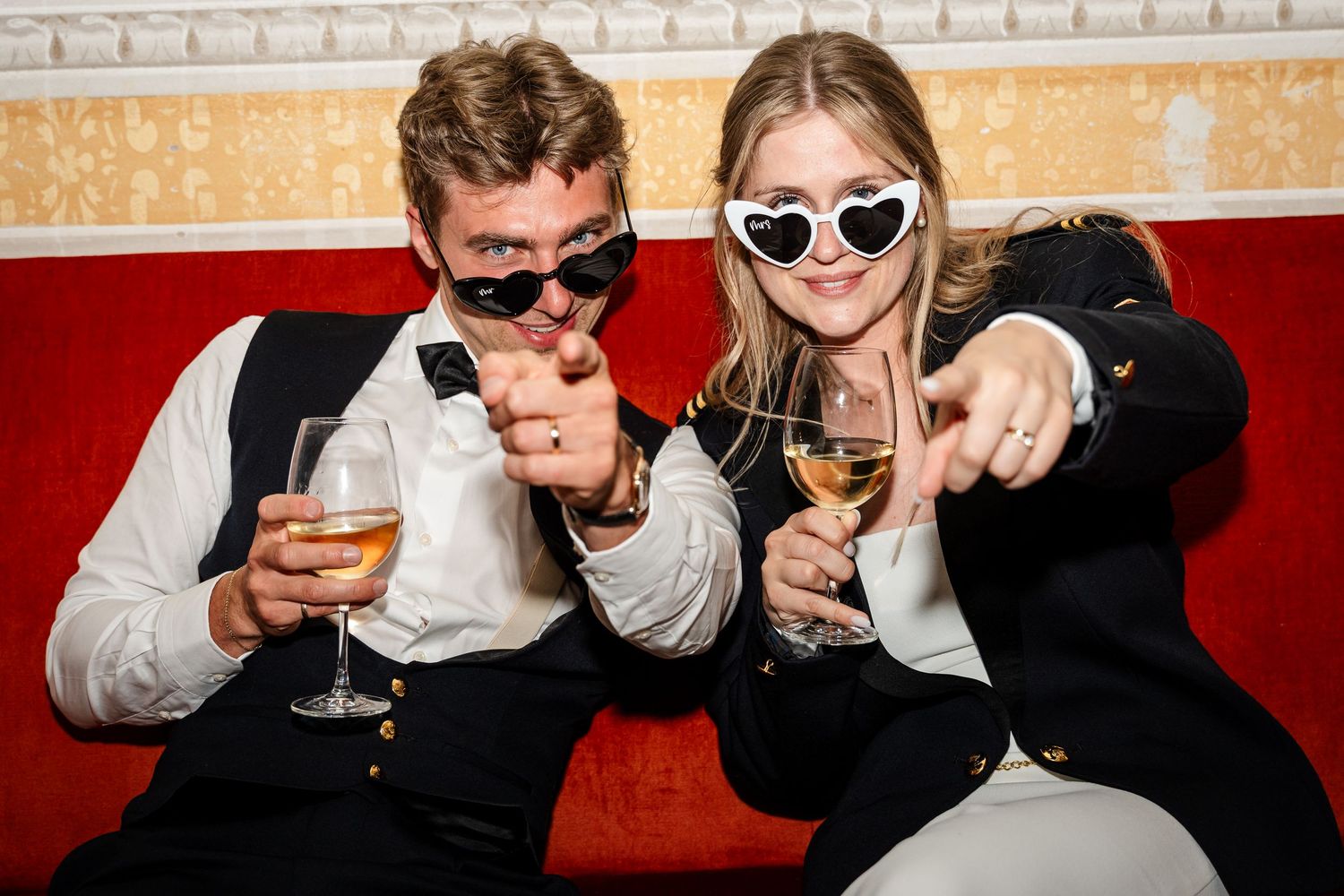 Two stylish young adults wearing sunglasses point at camera while holding wine glasses on a red sofa.