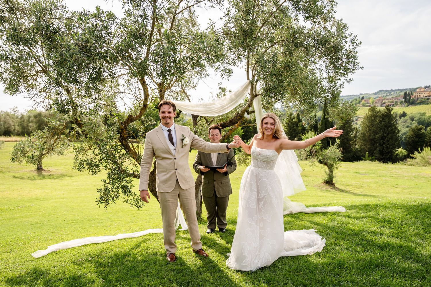 Joyful bride and groom celebrate after outdoor wedding ceremony beneath olive trees on lush green lawn.