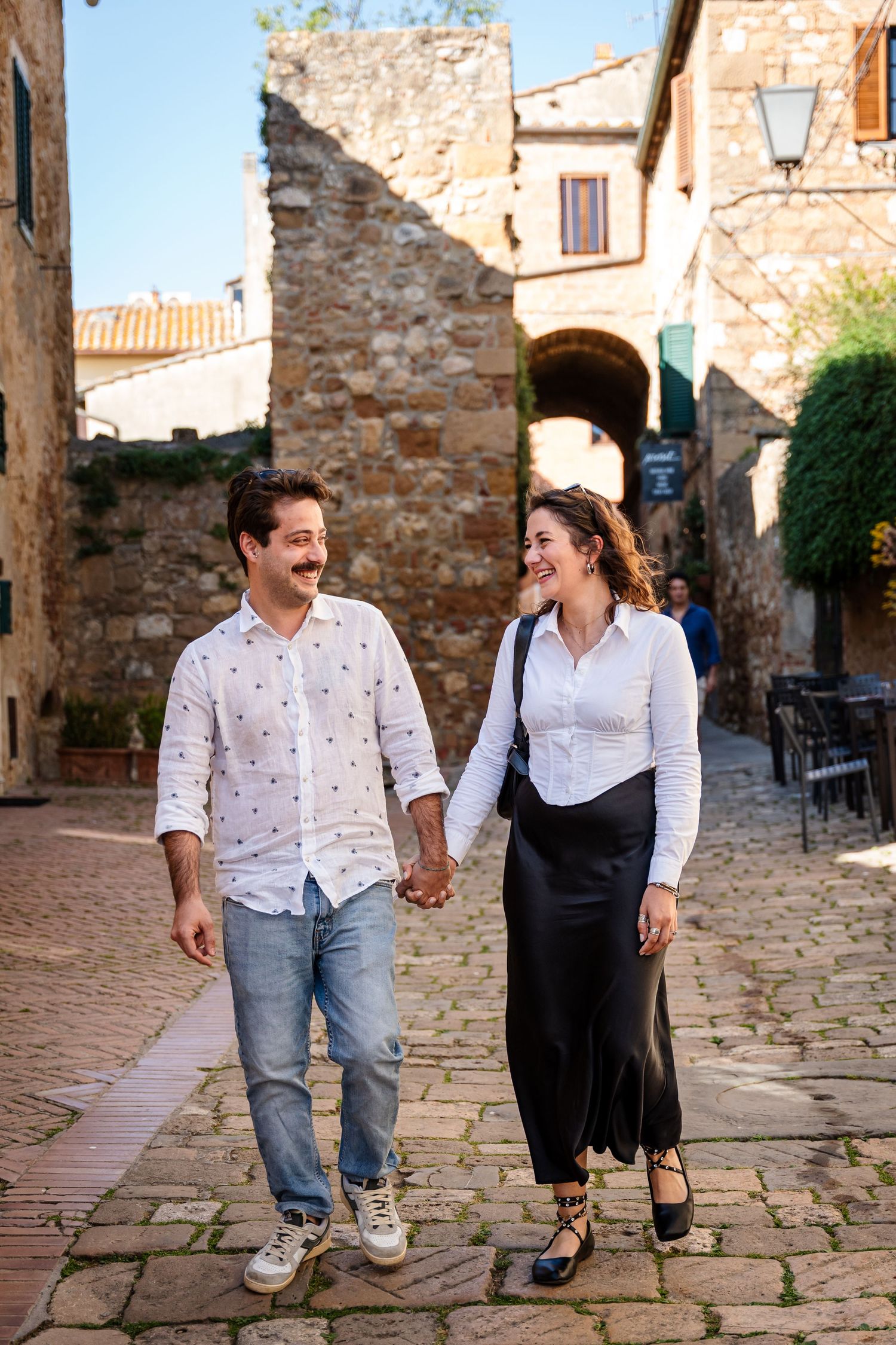 Couple holding hands and smiling while walking on cobblestone street in a historic Tuscan village on a sunny day.