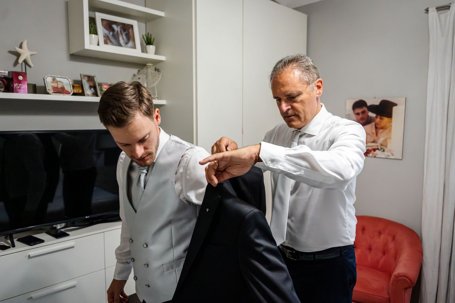 Older man helping young groom put on suit jacket indoors before wedding ceremony.
