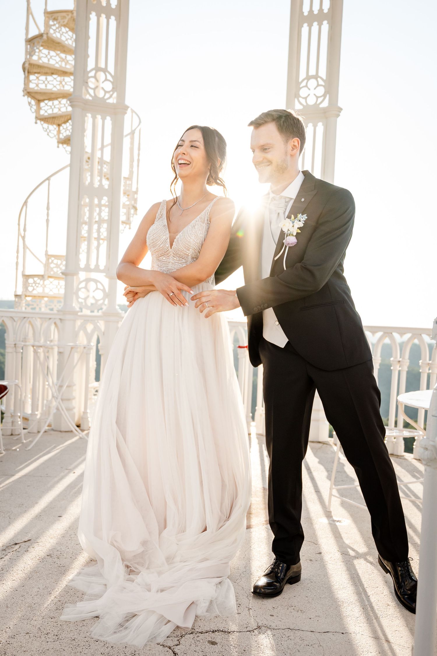 Bride and groom laughing together on a sunlit rooftop terrace, bride in flowing white gown, groom in black tuxedo.