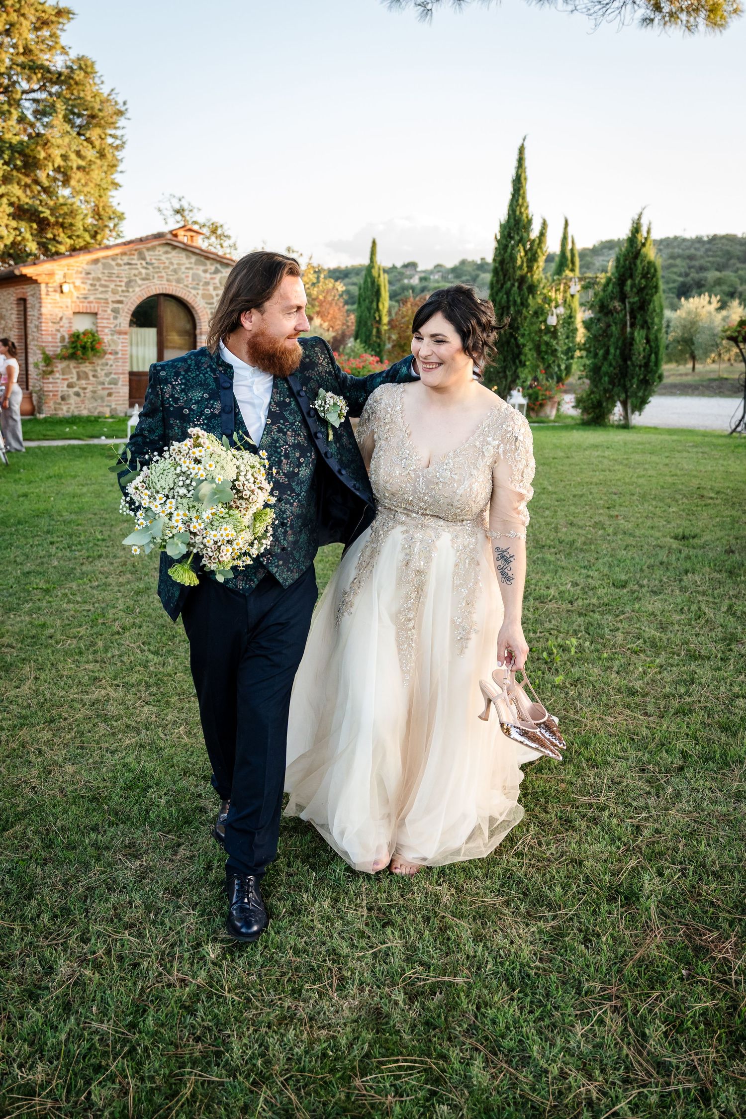 A couple in wedding attire share a romantic moment on a grassy lawn with cypress trees and stone buildings in the background.