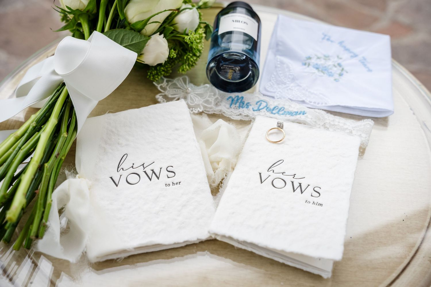 Two white vow books labeled his and her vows displayed on a table with wedding rings, bouquet, and blue ink bottle.