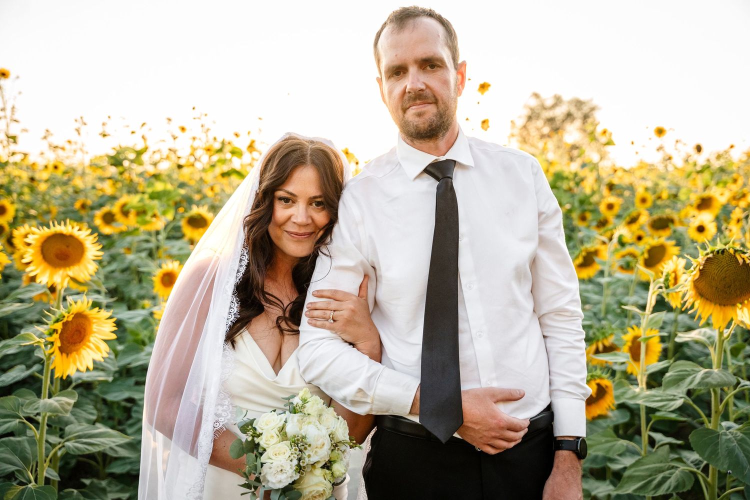 Bride and groom pose together smiling in a vibrant sunflower field at sunset.