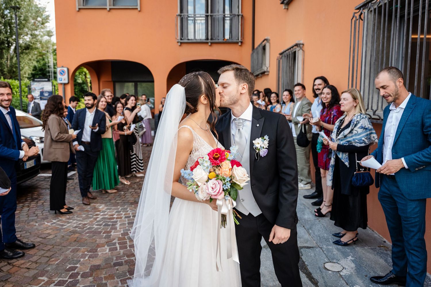 Newlyweds sharing first kiss outside while guests celebrate.